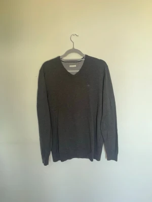 Mörkgrå V-ringad overshirt/ Pullover TomTailor - Mörkgrå V-ringad overshirt/ pullover från TomTailor med diskret logga på bröstet. Tröjan har långa ärmar och ribbade muddar vid ärmslut och nederkant. Perfekt för lager på lager och enkel att matcha med olika stilar. Storlek M. Mycket fint skick