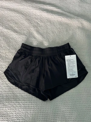 Svarta Hotty Hot shorts från Lululemon - Säljer ett par svarta Hotty Hot LR shorts från Lululemon. De har elastisk midja, meshpaneler, diskret dragkedjeficka och innerbyxa för extra täckning. Materialet är lätt, stretchigt och andas, perfekt för träning eller löpning.