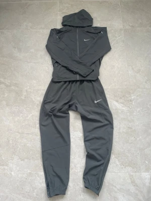 Mörkgrå Nikemillerset  - Snyggt mörkgrått träningsett från Nike med matchande jacka och byxor. Jackan har huva och dragkedja framtill, medan byxorna har resår vid benslut. Storlek: XS. Skick: Extremt fint! 
