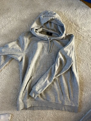 Grå hoodie med huva från H&M - En klassisk grå hoodie från H&M med stor huva och känguruficka framtill. Tillverkad i mjukt bomullsmaterial och har ribbade muddar vid ärmslut och nederkant. Perfekt för en avslappnad och chill stil. Lite mindre i storleken 