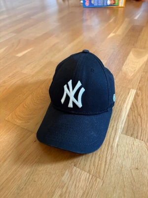 Svart New Era Yankees keps - Snygg svart keps från New Era med klassiska New York Yankees-loggan broderad i vitt framtill. Kepsen har böjd skärm och justerbar passform. Perfekt accessoar för en sportig och avslappnad stil.