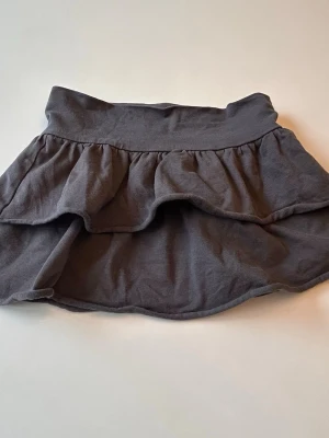Mörkgrå volangkjol i bomull - Supersöt mörkgrå kortkjol med volangdetalj. Kjolen har bred linning och är gjord i mjuk bomull som känns skön mot huden. Perfekt för dig som gillar en enkel men ändå trendig look.