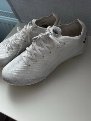  Nike fotbollsskor  - Säljer ett par stilrena vita fotbollsskor från Nike med klassisk snörning och låg profil. Skorna har en ovandel i textilmaterial med diskret struktur och en svart Nike-logga på hälen. Perfekta för dig som vill ha en lätt och smidig sko på planen. 