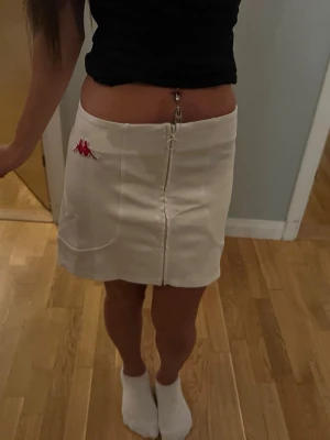 Vit kortkjol från Kappa med dragkedja - Säljer en vit kortkjol från Kappa med en snygg silverfärgad dragkedja framtill och Kappa-logga i rött på vänster sida. Kjolen har en enkel, rak passform och är tillverkad i ett mjukt material som känns bekvämt. Perfekt till sommaren!
