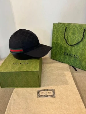 Svart keps med GG-mönster - Snygg svart keps med klassiskt GG-monogram över hela kepsen. På sidan finns en ikonisk grön och röd rand. Kepsen har böjd skärm och justerbar rem bak. Materialet är troligtvis bomullscanvas med detaljer i textil. Perfekt för dig som vill ha en exklusiv och trendig look.