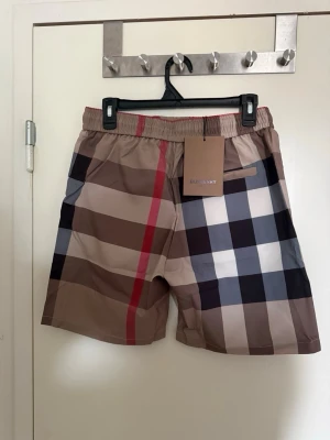 Burberry rutiga badbyxor beige - Säljer ett par snygga badbyxor från Burberry med klassiskt beige, svart, vitt och rött rutmönster. De har resår i midjan, svarta dragsnören och sidofickor. Materialet är snabbtorkande polyester, perfekt för stranden eller poolen.