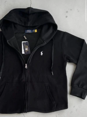Svart hoodie från Polo Ralph Lauren - Snygg svart hoodie från Polo Ralph Lauren med dragkedja framtill och klassisk vit logga på bröstet. Hoodien har huva med dragsko och två fickor fram. Tillverkad i mjukt bomullsmaterial som känns skönt mot huden. Perfekt för en avslappnad och stilren look.
