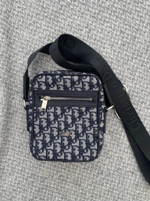 Mörkblå axelväska från Dior - Snygg axelväska från Dior i mörkblått och vitt monogramtyg med justerbar svart axelrem där det står 'Christian Dior'. Väskan har en fyrkantig form, en dragkedjeficka framtill och klassisk Dior-logga över hela tyget. Perfekt för dig som gillar designer och streetstyle.