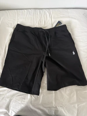Svara polo Ralph Lauren  - Snygga svarta  shorts  Polo Ralph Lauren med klassisk broderad logga i vitt på benet. Shortsen har elastisk midja med snörning och är tillverkade i mjuk bomull, perfekta för en avslappnad stil.