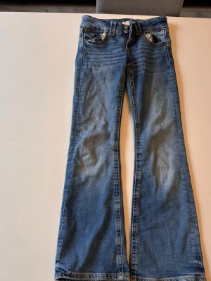 Blå bootcut jeans med låg midja - Säljer ett par blå bootcut jeans med låg midja och slitningar framtill. Jeansen har klassiska fem fickor, dubbla knappar i linningen och vida ben. Perfekta för en avslappnad och trendig look. Materialet är mjuk denim i bomull.