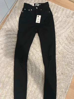 Svarta skinny jeans från 157 - Säljer ett par svarta skinny jeans från 157 med hög midja och slitningar vid knäna. Jeansen har klassisk femficksdesign, silverfärgade nitar och knapp, samt bälteshällor. Materialet är stretchigt för en bekväm passform.
