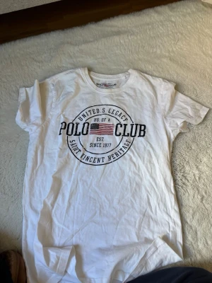 Vit t-shirt från Polo Club Saint Vincent - Vit t-shirt från Polo Club Saint Vincent med tryck framtill i svart och amerikansk flagga. Klassisk rund hals och korta ärmar. Skön och enkel modell i bomull, perfekt till jeans eller shorts.