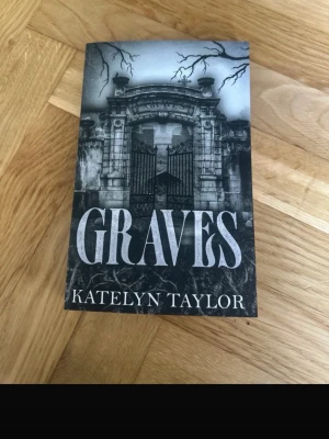Graves av Katelyn Taylor - En svartvit pocketbok med titeln 'Graves' av Katelyn Taylor. Omslaget har en mystisk port i sten och trädgrenar mot en dramatisk himmel. Boken har ett mörkt, gotiskt tema och är tryckt på vanligt papper. Perfekt för dig som gillar spänning och romantik.