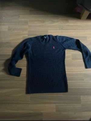 Mörkblå stickad tröja från Ralph Lauren - Säljer en mörkblå stickad tröja från Ralph Lauren med klassiskt kabelstickat mönster och rund halsringning. Tröjan har långa ärmar och en liten röd logga broderad på bröstet. Perfekt för dig som gillar stilrena och tidlösa plagg.