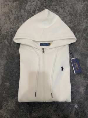 Vit hoodie från Polo Ralph Lauren - Snygg vit hoodie från Polo Ralph Lauren med dragkedja framtill och klassisk huva. Broderad marinblå logga på bröstet och ribbade muddar vid ärmar och nederkant. Tillverkad i mjuk bomullsblandning för skön känsla. Perfekt för en clean och stilren look.