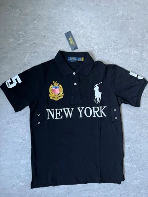Polo Ralph Lauren New York piké - Svart pikétröja från Polo Ralph Lauren med vit broderad logga på bröstet, stort 'NEW YORK'-tryck och guldfärgad patch. Tröjan har korta ärmar, krage och knappar framtill samt siffran 5 på ärmen. Perfekt för en sportig och stilren look.