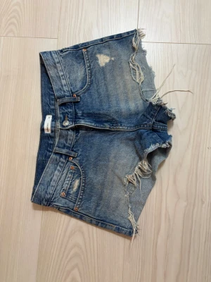 Blå jeansshorts med fransar - Säljer ett par blå jeansshorts från gina tricot med råa, fransiga kanter och slitna detaljer. Klassisk femficksmodell med knapp och dragkedja framtill. Perfekta för sommaren och ger en avslappnad vibe.