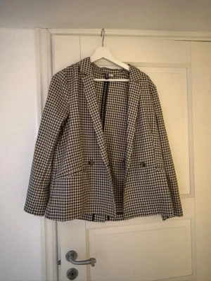 Rutig kavaj med dubbelknäppning - Snygg rutig kavaj i svart, vitt och beige med klassisk dubbelknäppning och två framfickor. Kavajen har en rak passform och smala slag. Perfekt för dig som vill ha en trendig och stilren look. Materialet känns mjukt och följsamt.