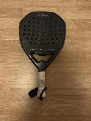 Svart padelracket från Xcalion - Padelracket från Xcalion i svart med vit lindad greppyta. Racketen har rund form med många hål och handledsrem för extra säkerhet. Perfekt för dig som vill ta ditt padelspel till nästa nivå.