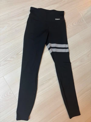 Svarta leggings från Stronger XS - Snygga svarta leggings från Stronger i storlek XS. De har två vita ränder med meshdetalj på ena benet. Tillverkade i 73% återvunnen polyester och 27% elastan för en stretchig och bekväm passform. Perfekta för träning eller chill.