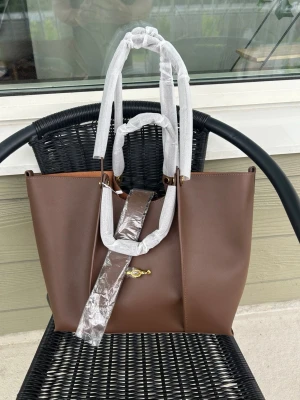 Brun tote handväska med gulddetalj - Snygg brun handväska i tote-modell med raka linjer och guldfärgad vridknäppe framtill. Väskan har dubbla handtag och är tillverkad i slätt konstläder. Perfekt för dig som gillar stilrena accessoarer med en modern touch.