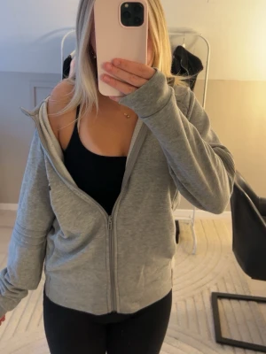 Grå zip hoodie från Adidas - Säljer en grå hoodie från Adidas med dragkedja och svart Adidas-logga på ena sidan. Hoodien har huva och passar till träning men även som mjukiskläder. 