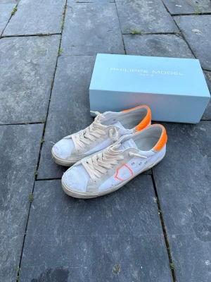 Philippe Model vita sneakers med orange detalj - Säljer ett par vita Philippe Model sneakers med orange häl och orangea loggan på sidan samt orangea detaljer längst bak på skon. Skorna har klassisk låg profil, vita snören och sula i gummi. Materialet är en mix av läder och textil.