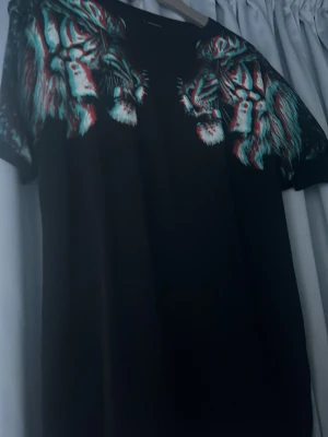 Marcelo burlon T-shirt  - Cool svart t-shirt med stort, färgstarkt tigertryck över axlar och bröst. Trycket har en 3D-effekt i rött, blått och vitt som verkligen sticker ut. Perfekt för dig som vill ha en unik och edgy stil. Klassisk passform och rund hals.