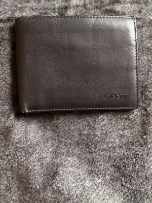 Svart läderplånbok från GANT - Stilren svart plånbok i äkta läder från GANT. Klassisk vikbar modell med flera kortfack på båda sidor och diskret logga präglad i hörnet. Perfekt för dig som vill ha en enkel och snygg accessoar i fickan.