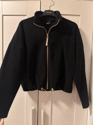 Svart fleecejacka från Gina Tricot - Mysig svart fleecejacka från Gina Tricot med dragkedja framtill och hög krage. Jackan har justerbar dragsko i nederkant och är tillverkad i mjukt fleecematerial. Perfekt för lager på lager och enkel att matcha med olika outfits.