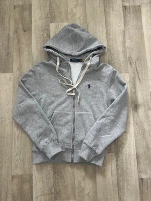 Grå hoodie från Polo Ralph Lauren - Snygg grå hoodie från Polo Ralph Lauren med dragkedja, huva och vita snören. Klassisk broderad logga på bröstet och känguruficka framtill. Perfekt för en chill och avslappnad stil.