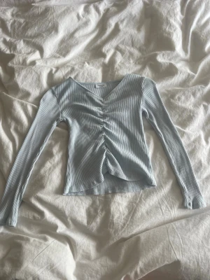 Ljusblå ribbad topp med snörning - Trendig ljusblå långärmad topp med ribbad struktur och snygg snörning framtill som ger en rynkad effekt. V-ringad halsringning och figurnära passform. Perfekt att styla med jeans eller kjol för en cool look.