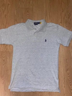 Grå pikétröja från Polo Ralph Lauren - Klassisk ljusgrå pikétröja från Polo Ralph Lauren med korta ärmar och krage. Tröjan har två knappar vid halsen och den ikoniska broderade loggan i marinblått på bröstet. Tillverkad i mjuk bomull för en bekväm känsla.
