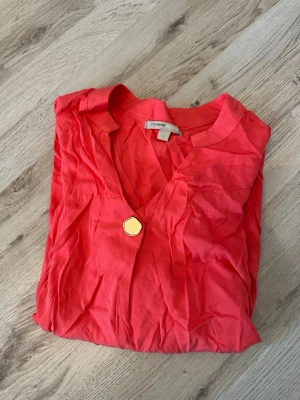 Röd blus från MyWear med knapp - Snygg röd blus från MyWear i lätt och mjukt tyg. Blusen har V-ringning och en dekorativ guldig knapp framtill. Perfekt till jeans eller kjol för en trendig look. Kortärmad modell som ger en avslappnad och modern känsla.