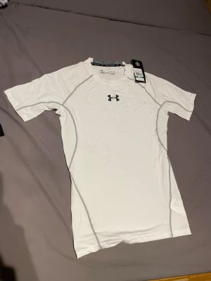 Vit Under Armour tränings-tshirt - Säljer en vit tränings-tshirt från Under Armour med grå sömmar och logga på både bröst och rygg. Materialet är stretchigt och fuktavvisande, perfekt för intensiva träningspass. Kortärmad och figurnära passform som ger bra rörelsefrihet.