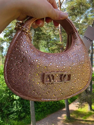 Glittrig guld-rosa handväska  - Super snygg väska, den är mycket snyggare i verkligheten. Den har lång axelrem också. Väskan är ett rent konstverk, helt täckt av ett tätt hav av gnistrande strasskristaller i olika storlekar. Kristallerna fångar och reflekterar ljuset i varje vinkel, vilket skapar en magisk, tredimensionell skimmereffekt.  Storleken: Längd: 20 cm Höjd: 20 cm Bredd/Djup: 8 cm
