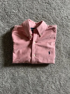 Rosa skjorta från Ralph Lauren - Klassisk rosa skjorta från Ralph Lauren med button-down krage och broderad logga på bröstet.  Storlek  vilket motsvarar medium ganska bra. Mycket bra skick!