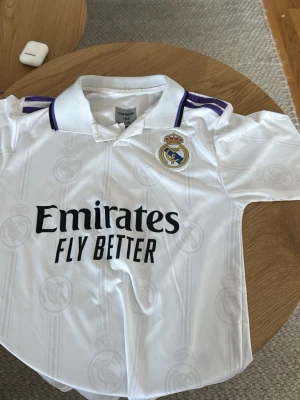 Real Madrid matchtröja Benzema - Säljer en vit Real Madrid matchtröja från Adidas med klubbmärke, sponsortryck och lila detaljer på axlarna. På ryggen står det Benzema och nummer 9. Tröjan har krage och är tillverkad i ett lätt, ventilerande material.