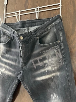 Svarta Dsquared2 ICON jeans - Säljer ett par svarta Dsquared2 ICON jeans med slitningar och tryck på vänster ficka. Jeansen har smal passform, klassisk femficksdesign och coola detaljer som ger en streetwear vibe. Materialet är denim med stretch för extra komfort.