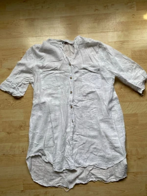 Vit linneskjorta med knappar - Luftig vit blus i linne med knappar framtill och två bröstfickor. Blusen har trekvartsärm och en avslappnad passform med rundad nederkant. Perfekt för en chill och stilren look.