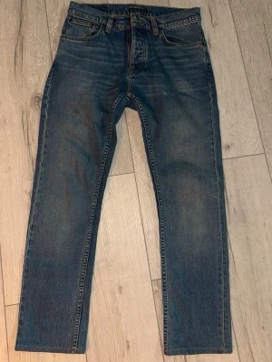 Nudie Jeans Grim Tim True Gold 30/32 - Snygga blå Nudie Jeans i modellen grim tim.  Klassisk femficksdesign, de har slimfit passform. Tillverkade i 99% bomull och 1% elastan för lite stretch. Perfekta för dig som gillar kläder som sitter bra och har en bra passform. De är i mycket bra skick, endast tvättats några gånger. Skriv till mig om ni undrar något😆