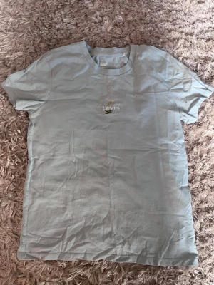Ljusblå Levi's t-shirt med broderi - Snygg ljusblå t-shirt från Levi's med ett broderat logotypmärke och blommotiv på bröstet. Klassisk rund hals och korta ärmar. Tillverkad i mjuk bomull som känns skön mot huden. Perfekt för en avslappnad och trendig look.