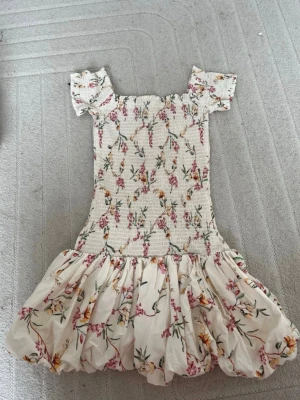  Trendig slutsåld Blommig kortklänning med volang - Supersöt kortklänning med blommigt mönster i rosa, gult och grönt på vit botten. Klänningen har puffiga korta ärmar, smockad överdel och volangig kjol. Perfekt för sommarens alla tillfällen!
