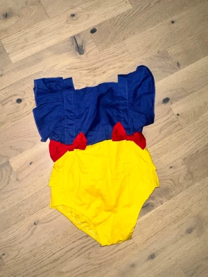 Snövit-inspirerad maskeraddräkt barn - Söt maskeraddräkt för barn inspirerad av Snövit, med blå överdel och gula volangprydda shorts. Dräkten har röda rosetter framtill och knytband i ryggen. Tillverkad i mjukt tyg, perfekt för maskerad eller cosplay.