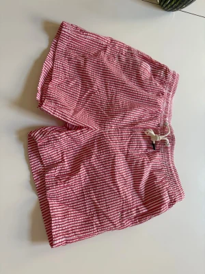 Badshorts. Randiga röda snörning - Säljer ett par röda och vita randiga badshorts med elastisk midja och vit snörning. Shortsen har en avslappnad passform och är perfekta för varma dagar. Tillverkade i ett lätt och bekvämt material som känns skönt mot huden.