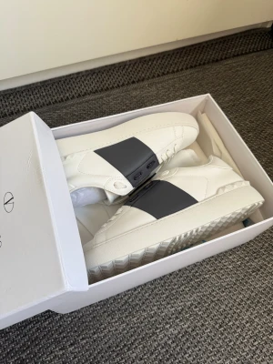 Valentino vita sneakers med svart band - Snygga vita sneakers från Valentino Garavani med ett brett svart band över vristen och coola nitar på sulan. Skorna har klassisk låg profil, vita skosnören och detaljer i läder. Perfekta för dig som vill ha en clean men ändå unik look.