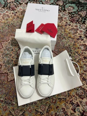 Valentino Garavani vita sneakers - Snygga vita sneakers från Valentino Garavani med bred svart rem över vristen och diskret logga på tungan. Skorna har klassisk låg profil, vita snören och är tillverkade i slätt läder. Perfekta för dig som gillar stilrena och exklusiva detaljer.