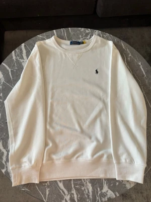 Vit sweatshirt från Polo Ralph Lauren - Vit sweatshirt från Polo Ralph Lauren med rund hals och broderad logga på bröstet. Lite större vid halsen och därför passar perfekt som en pullover på t.ex en skjorta eller en piké nu under sommarn.