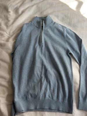 Ljusblå halvzip-tröja från Bläck - Snygg ljusblå tröja med halv dragkedja från Black. Tröjan har hög krage och är tillverkad i ett mjukt, tunt stickat material. Perfekt för lager på lager och enkel att matcha med olika outfits. Nypris: 699kr