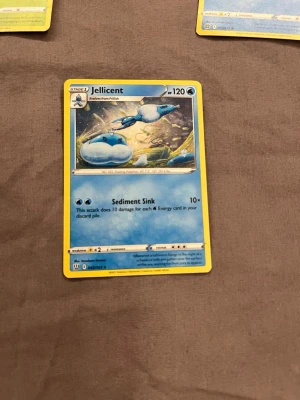 Pokémonkort Jellicent - Säljer ett Pokémonkort med Jellicent, en vatten-typ Pokémon. Kortet har blå och vita färger med en fantasifull illustration. Det är tillverkat i papper och har attacken Sediment Sink. Perfekt för samlare eller Pokémon-fans.